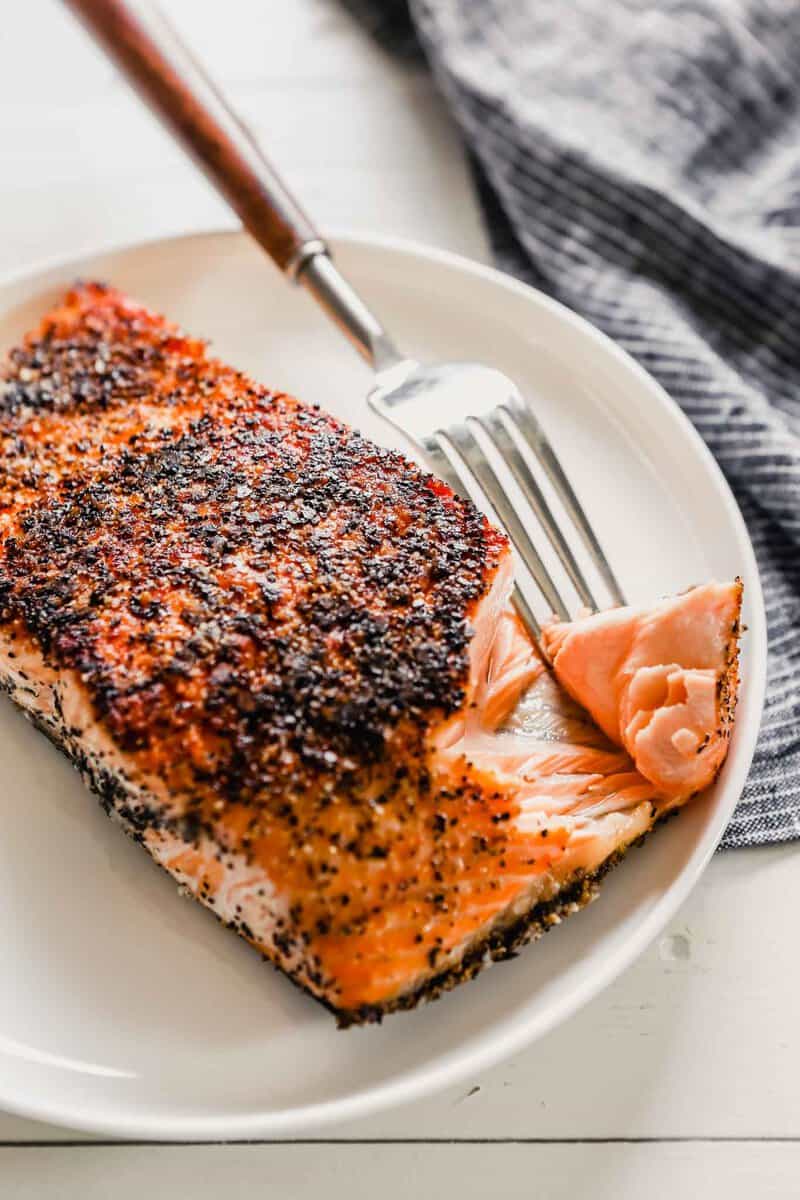 Close up photo of crispy pan seared salmon with skin set on a white plate with a fork taking a large chunk out of it