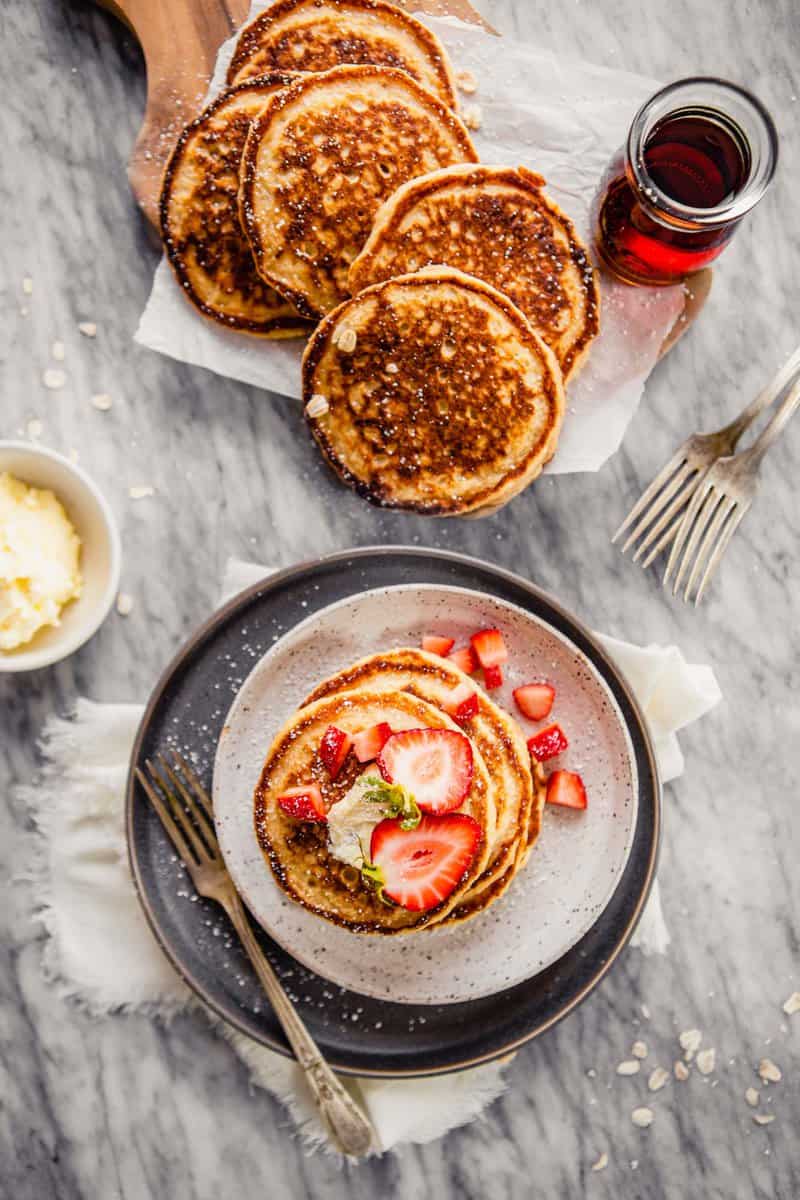 Overhead photo of pancakes stacked on a plate and pancakes arranged on a wooden platter with butter and maple syrup off to the side.
