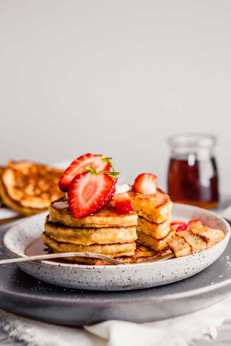 Photo of a bite take out of a stack of toasted oat pancakes on a plate topped with butter, strawberries and maple syrup