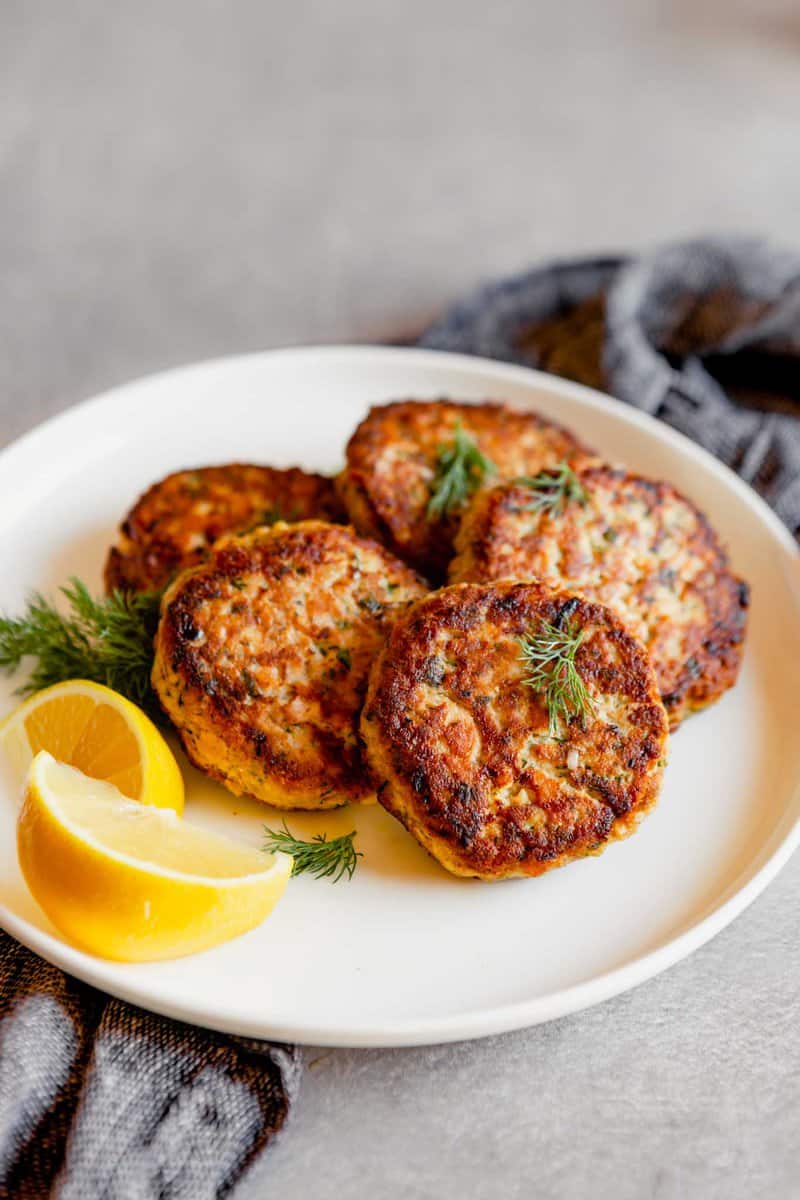 Photo of salmon cakes stacked on a white plate with lemon slices on a gray table with a blue napkin.