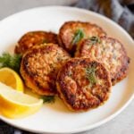 Close up photo of salmon cakes stacked on a white plate with lemon slices on a gray table with a blue napkin.