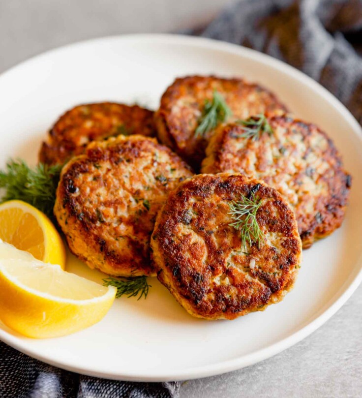 Close up photo of salmon cakes stacked on a white plate with lemon slices on a gray table with a blue napkin.