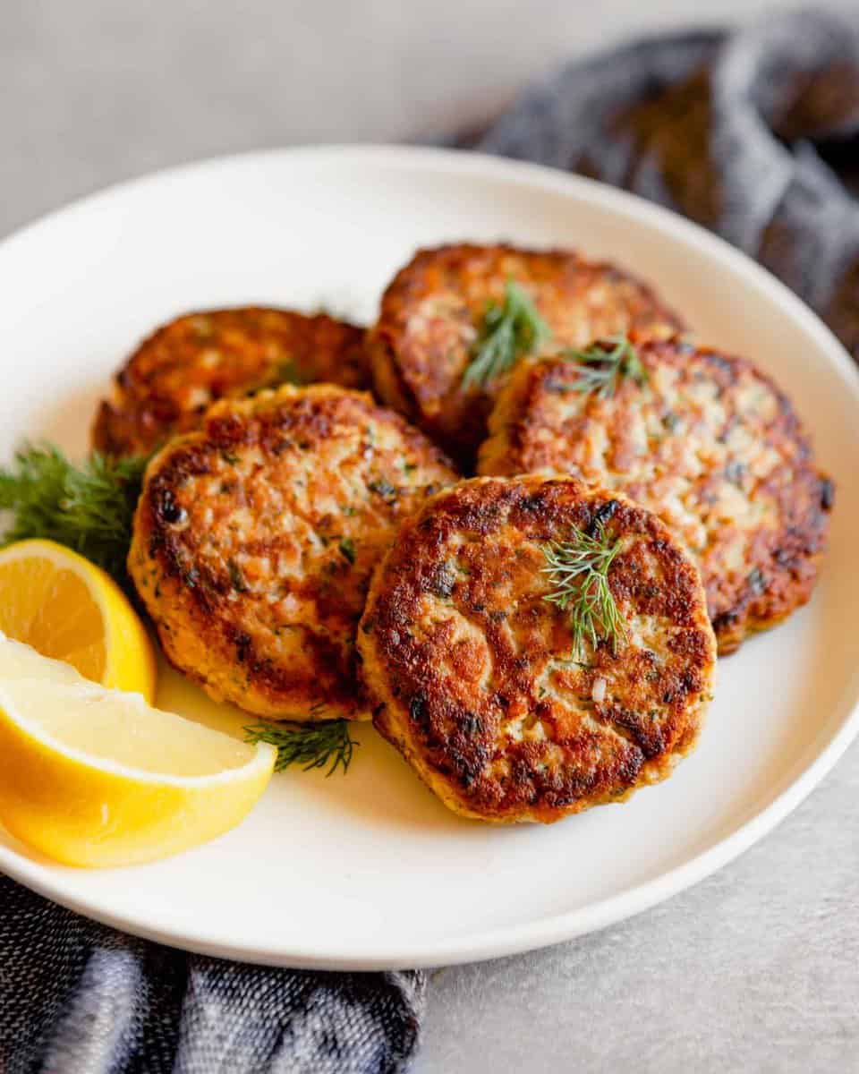 Close up photo of salmon cakes stacked on a white plate with lemon slices on a gray table with a blue napkin.