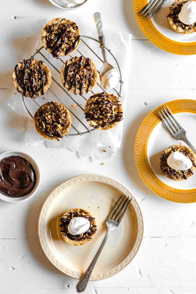 Overhead image of mini pecan pies (pecan tassies) arranged on a cooling rack and a varity of gold-colored plates on a white table