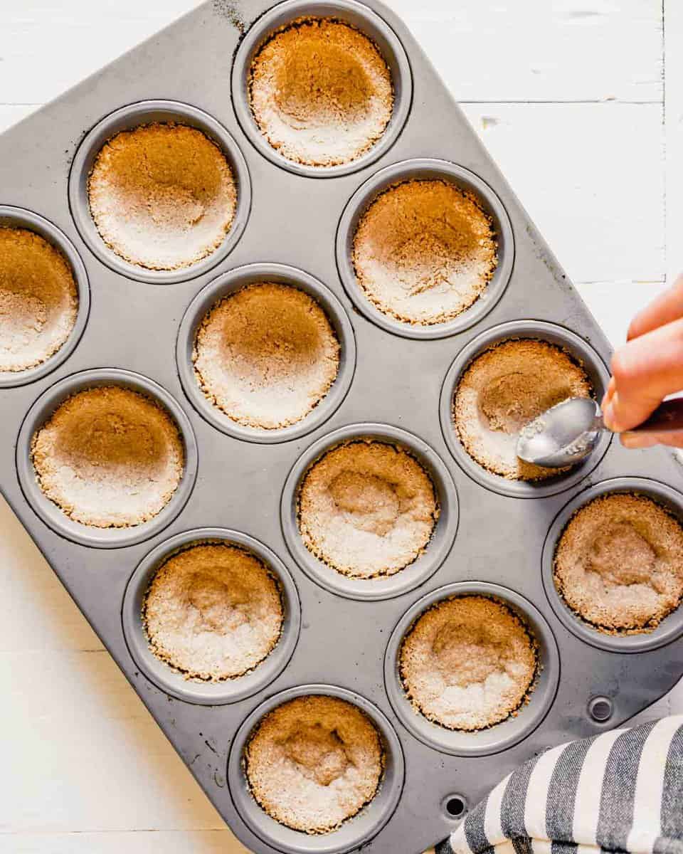 photo of someone pressing a spoon into baked mini crusts in a muffin tin, pressing them down after they have puffed up during baking