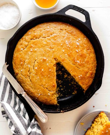 Overhead image of healthy cornbread in a cast iron skillet set on a wooden white table with a striped napkin and a knife