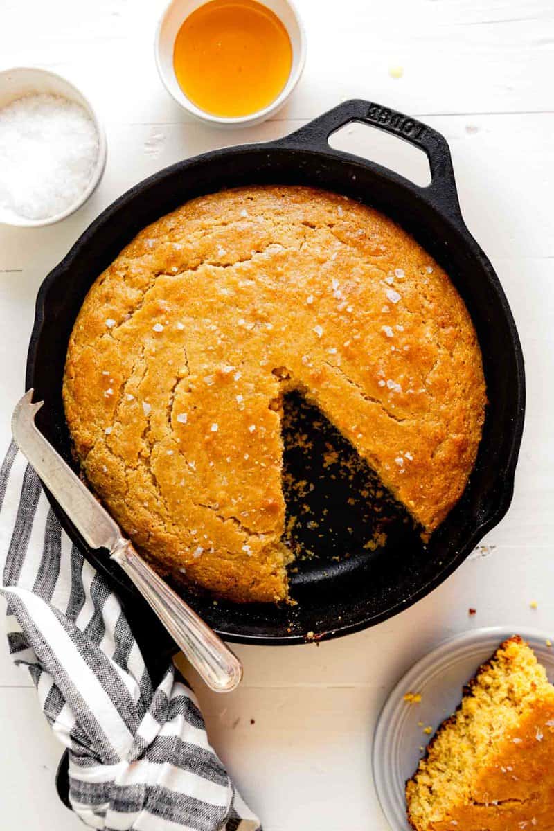 Overhead image of healthy cornbread in a cast iron skillet set on a wooden white table with a striped napkin and a knife