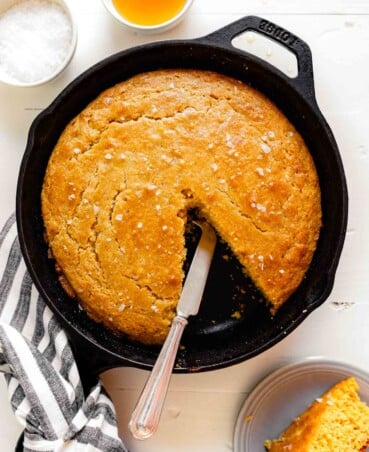Overhead image of healthy cornbread in a cast iron skillet set on a wooden white table with a bowl of salt and honey to the top right
