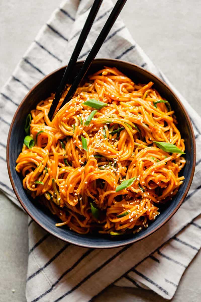 Overhead image of bright orange noodles with gochujang sauce in a dark blue bowl set on a striped napkin.