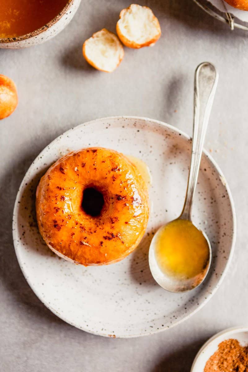 Overhead image of a glazed mochi donut on a white speckled plate with a spoon of glaze set off to the side. 