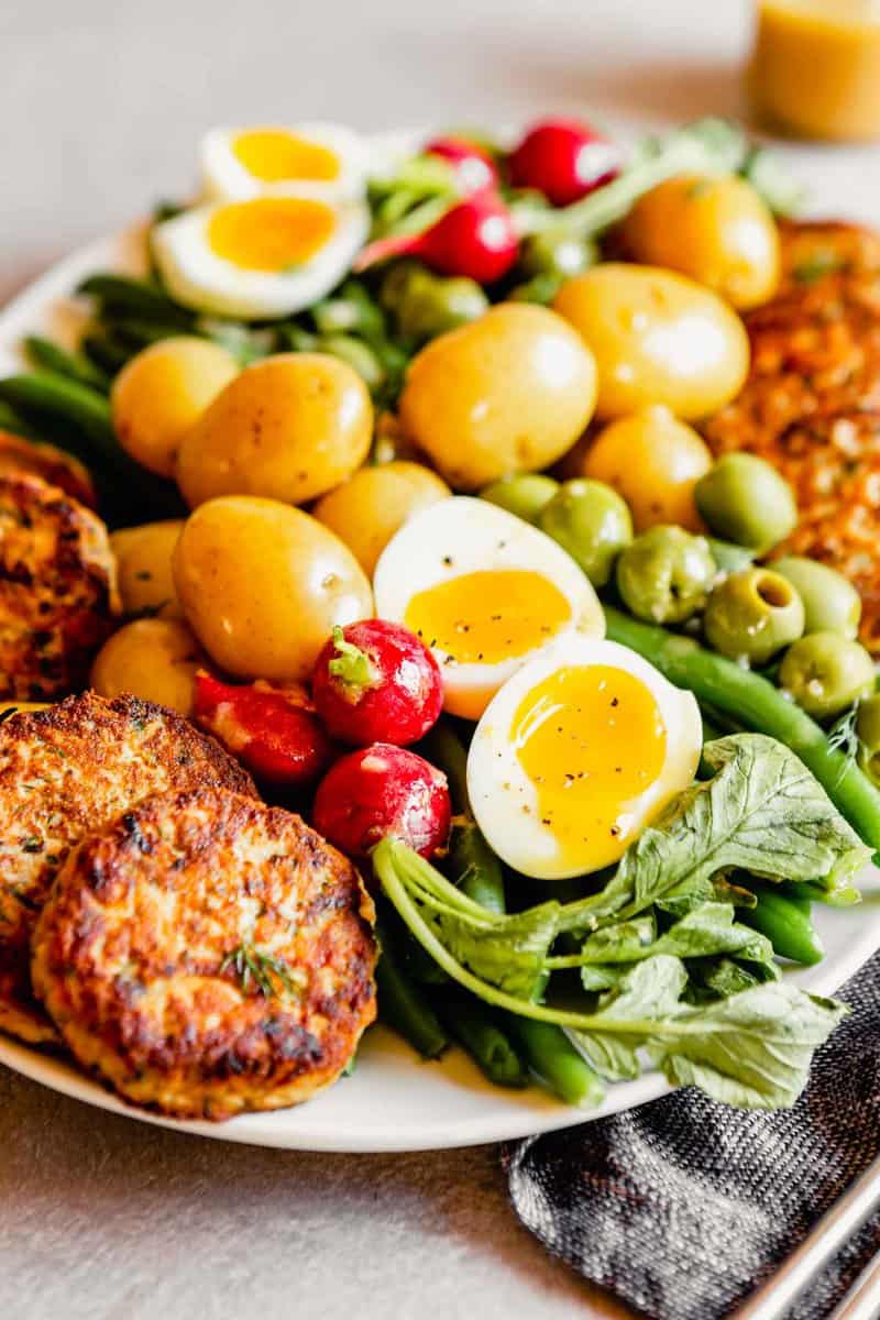 Side angle of a nicoise salad on a white oval plate. Focused on soft boiled eggs and salmon patties.