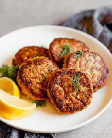 A white plate holds five golden-brown salmon patties garnished with dill, showcasing a classic salmon patties recipe. Two lemon wedges rest beside the patties, inviting a citrusy touch. A textured, dark-colored cloth is partially visible underneath the plate.