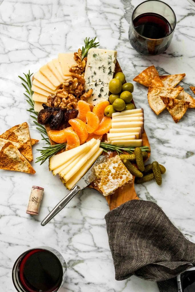 overhead image of a wood cutting board piled high with cheese, olives, clementines, rosemary and honeycomb