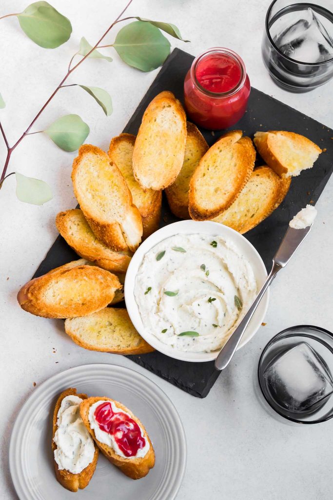overhead image of a slate board topped with a bowl of whipped ricotta and toasted crostini. A plate with mini toasts topped with ricotta and curd off to the side 
