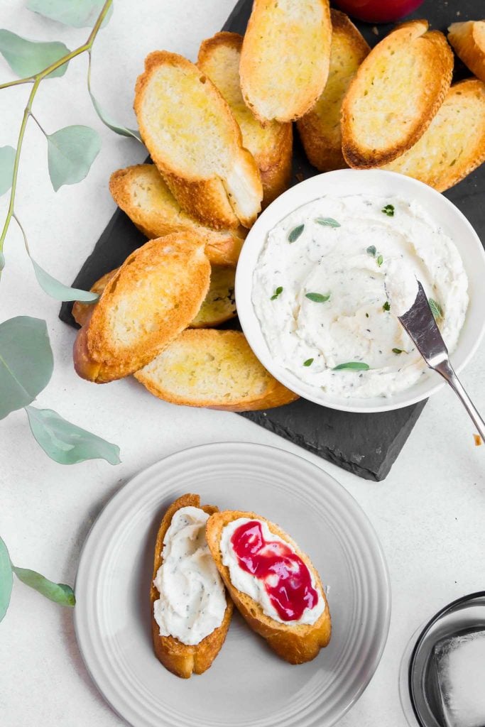 overhead image of a slate board topped with a bowl of whipped ricotta and toasted crostini. A plate with mini toasts topped with ricotta and curd off to the side 
