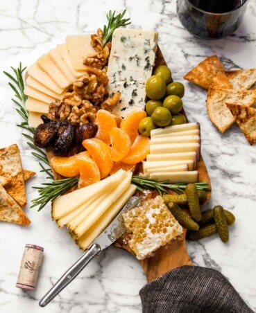 overhead image of a wood cutting board piled high with cheese, olives, clementines, rosemary and honeycomb