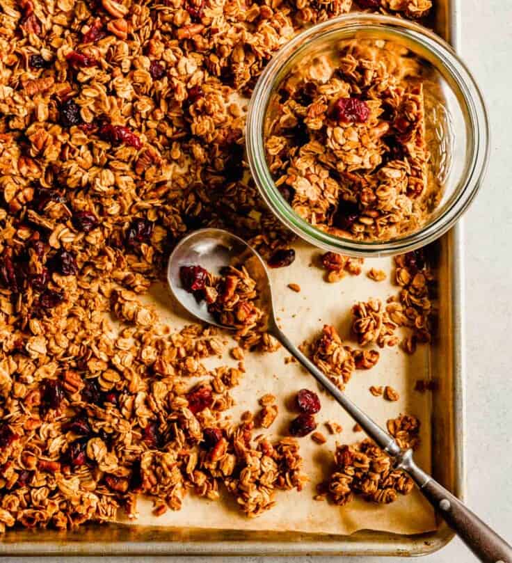 Overhead image of granola on a baking sheet with a spoon laid over top and a jar off to the side with granola in it