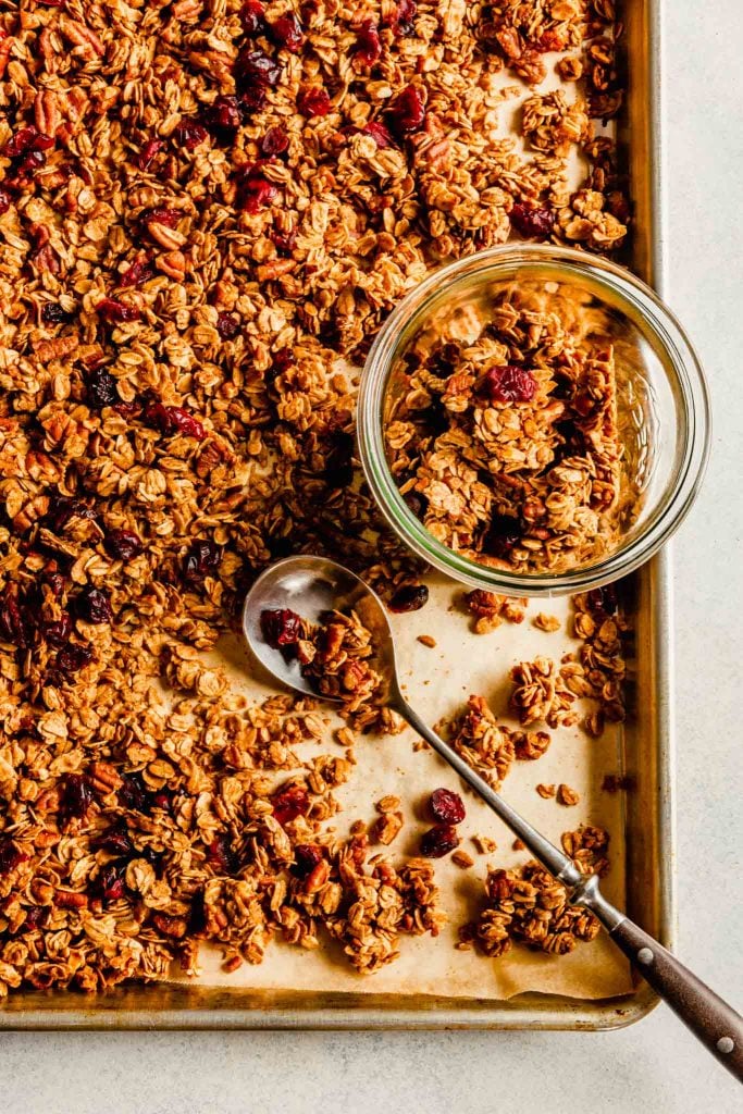 Overhead image of granola on a baking sheet with a spoon laid over top and a jar off to the side with granola in it