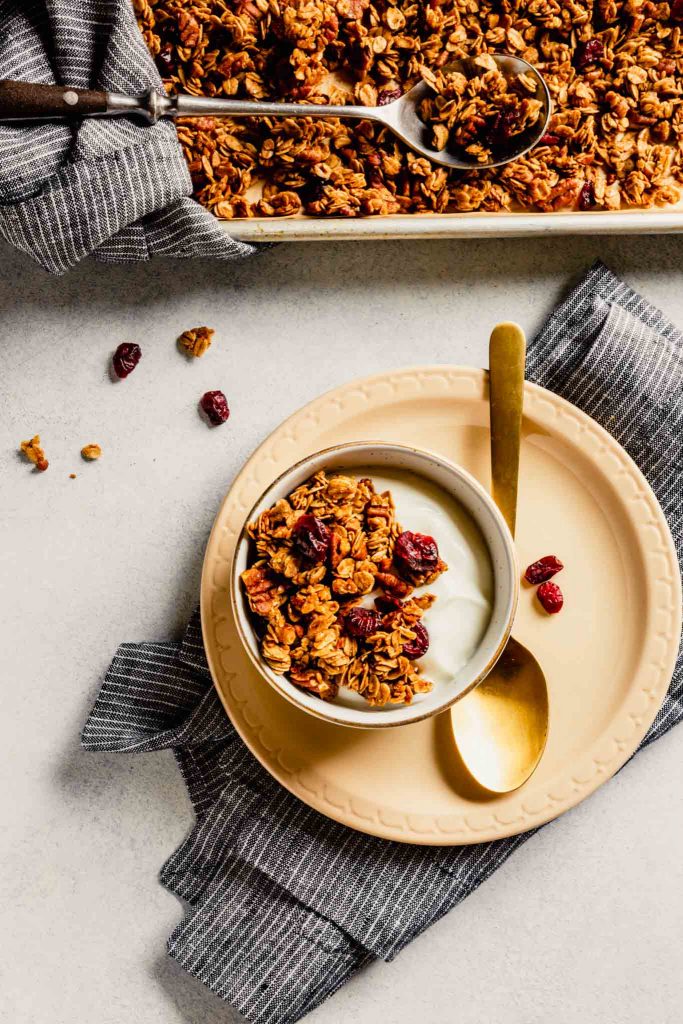 Overhead image of a white bowl filled with yogurt and granola on a cream colored plate with a baking sheet of granola in the background.