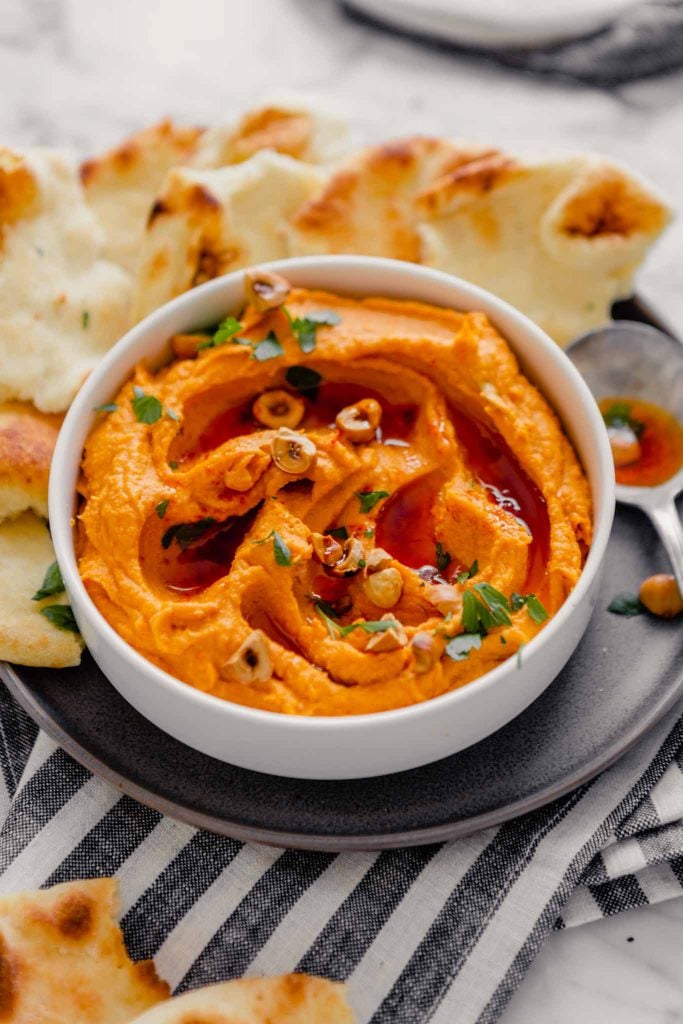 Side angle of orange hummus spread into a white bowl set on a gray plate on a marble table with fresh naan bread.