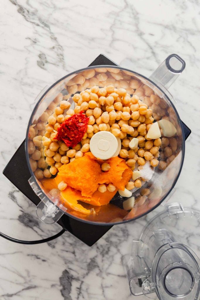 Overhead image of sweet potato puree, chickpeas, harissa and garlic in a food processor. 