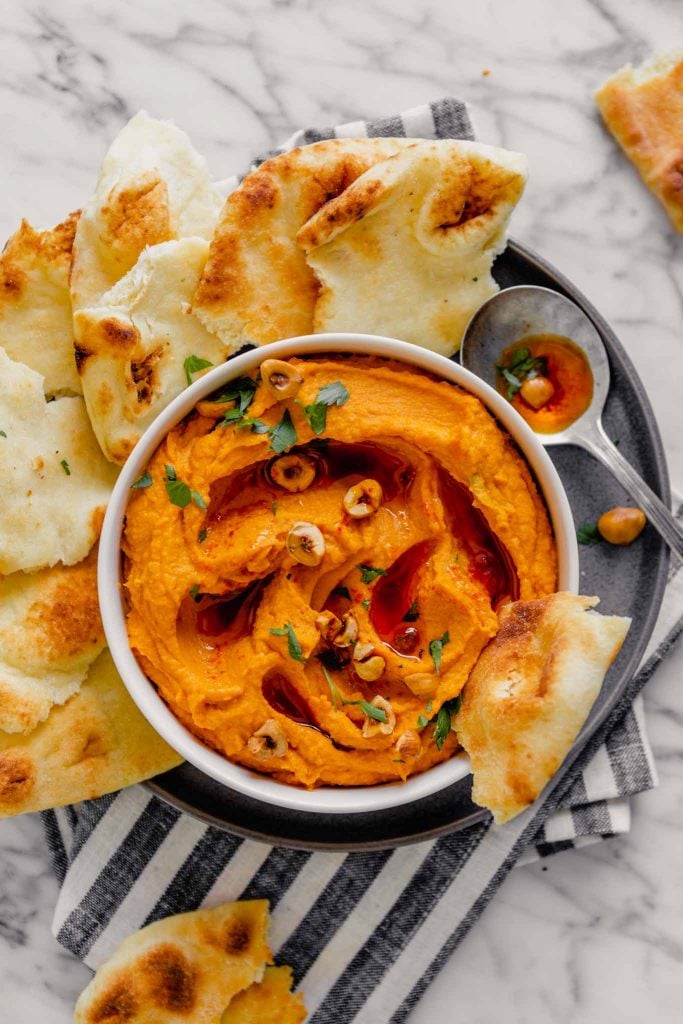 Overhead image of orange hummus spread into a white bowl set on a gray plate on a marble table with fresh naan bread.