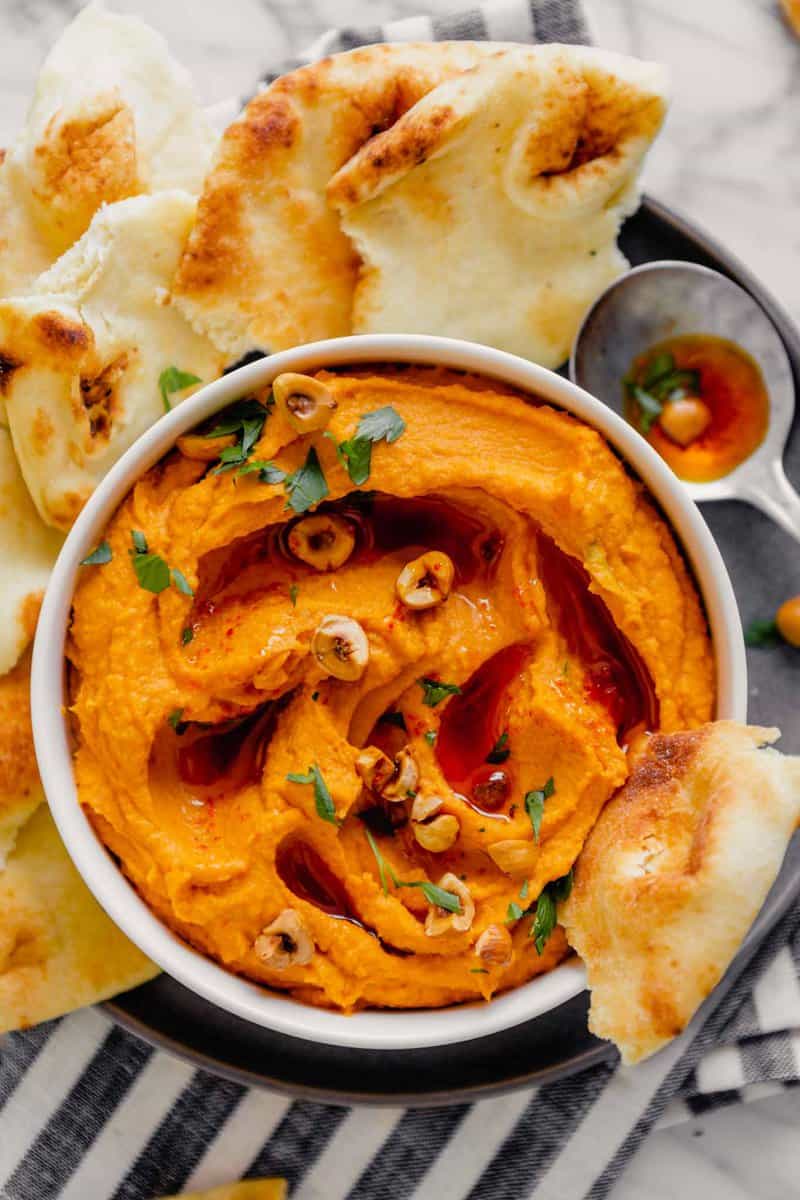 Overhead image of orange hummus spread into a white bowl set on a gray plate on a marble table with fresh naan bread.