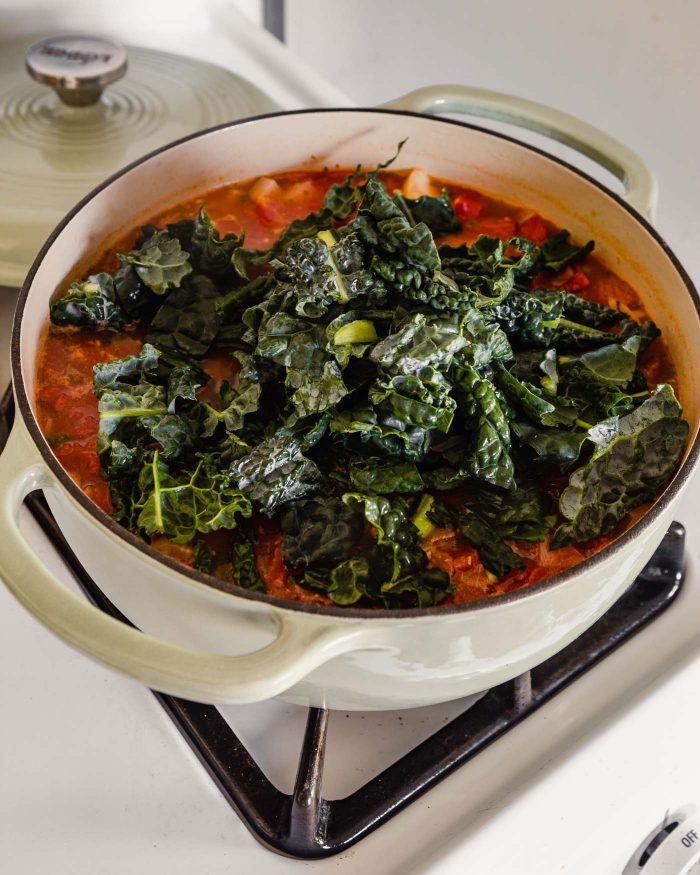 Heaping kale on top of a tomato soup mixture in a large Dutch oven on stove top