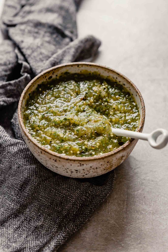 Side angle of green salsa in a white speckled bowl set on a gray table with a dark napkin off to the side