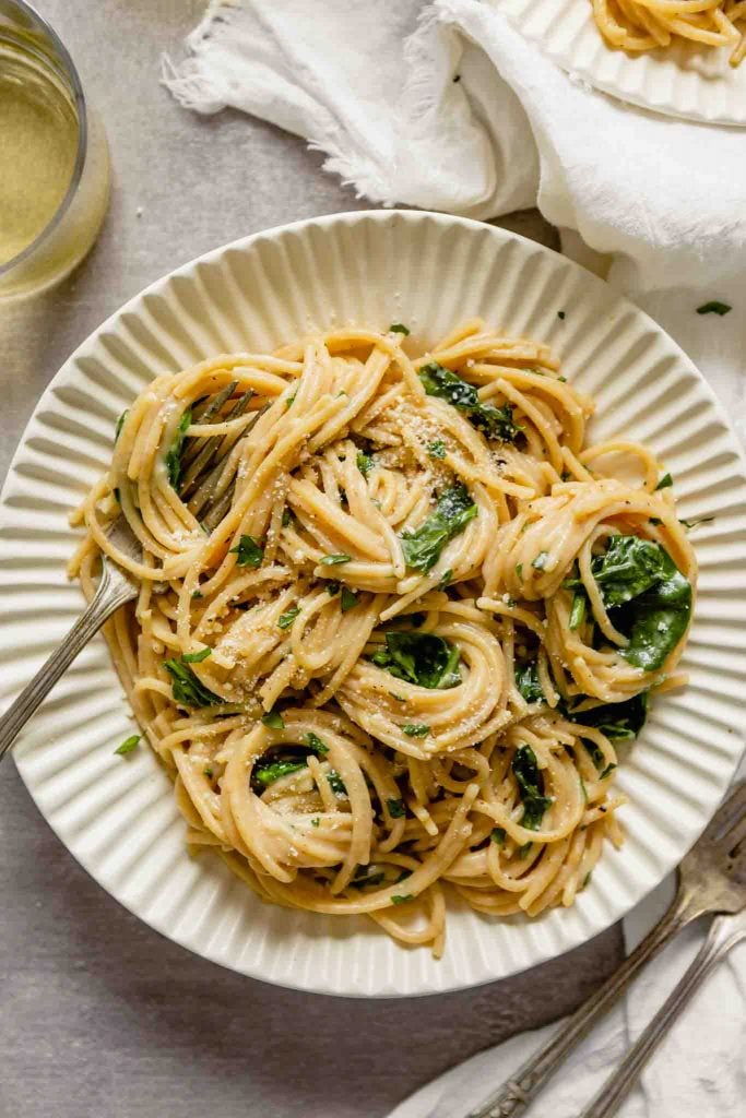 close up image of whole wheat pasta with spinach and Parmesan served in a white bowl with wine and parmesan off to the side