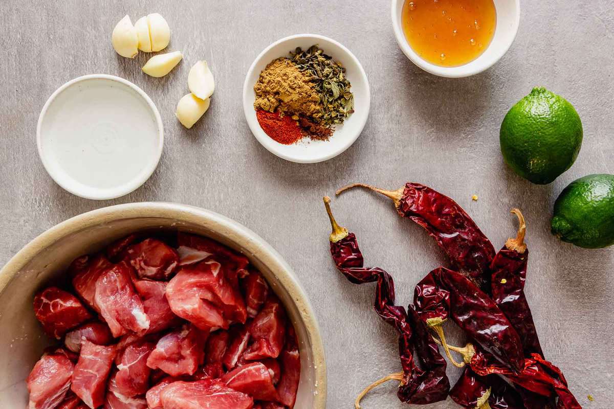 Overhead image of spices, dried chiles, pork, garlic and vinegar arranged on a gray table.