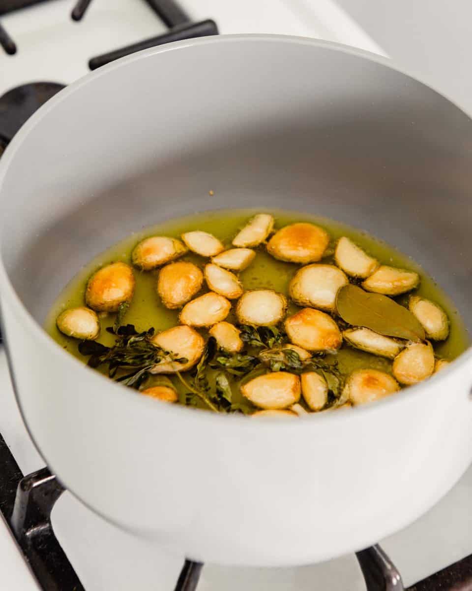 photo of lightly golden brown garlic cloves in a saucepan with oil and herbs