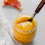 Side angle of orange curd in a small glass jar with a spoon set inside