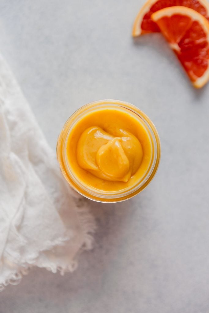 Close up overhead image of orange-colored curd in a glass jar set on a light blue table with a spoon and napkin off to the side