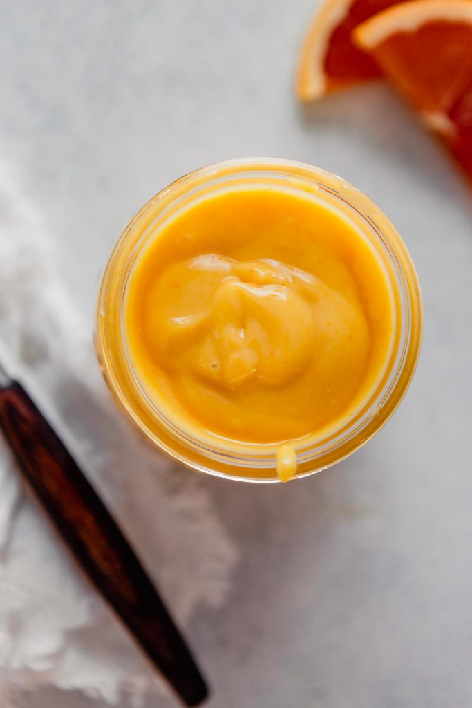 Close up overhead image of orange-colored  curd in a glass jar set on a light blue table with a spoon and napkin off to the side