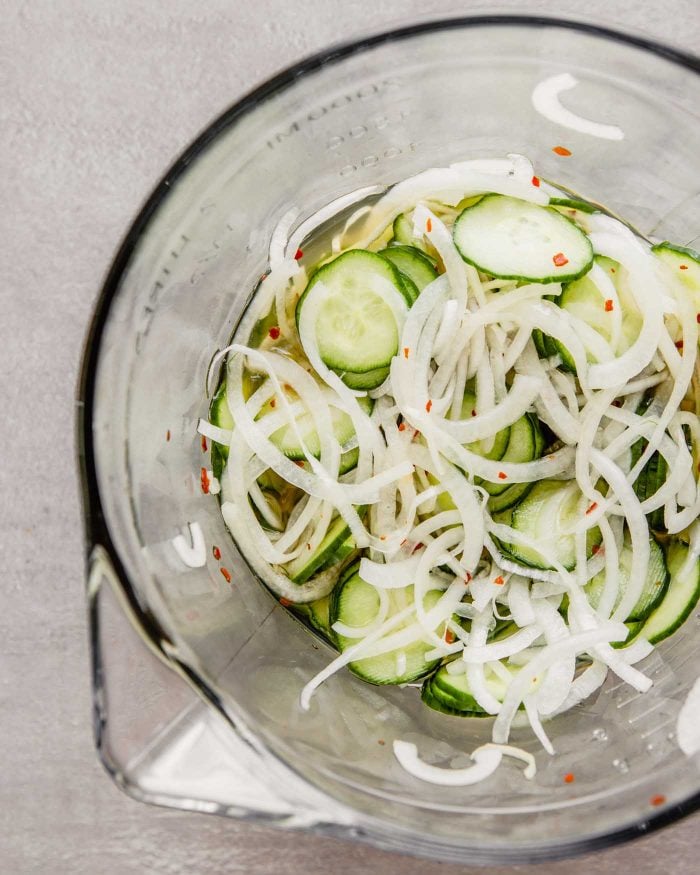 overhead image of thinly sliced cucumbers and onions in a glass bowl with a pickling liquid and red pepper flakes