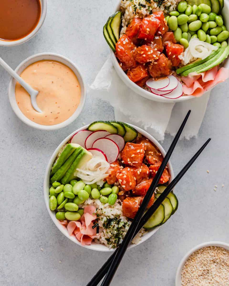 Overhead image of two bowsl filled with raw salmon, edamame, cucumbers, ginger and avocado set on a blue table with bowls of sauce and chopsticks