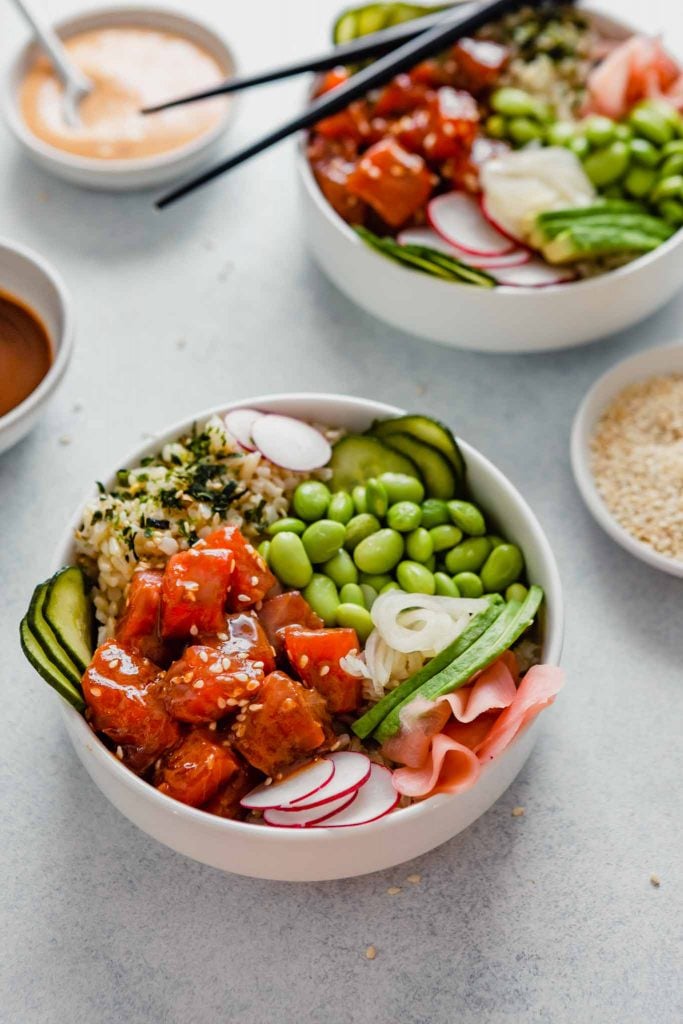 Side angle of a bowl filled with raw salmon, edamame, cucumbers, ginger and avocado set on a blue table with bowls of sauce and chopsticks