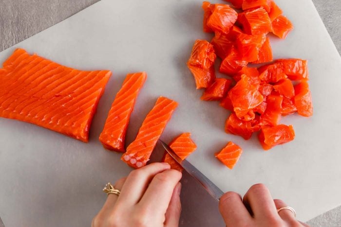 Overhead image of someone cutting raw salmon into cubes on a white cutting board.