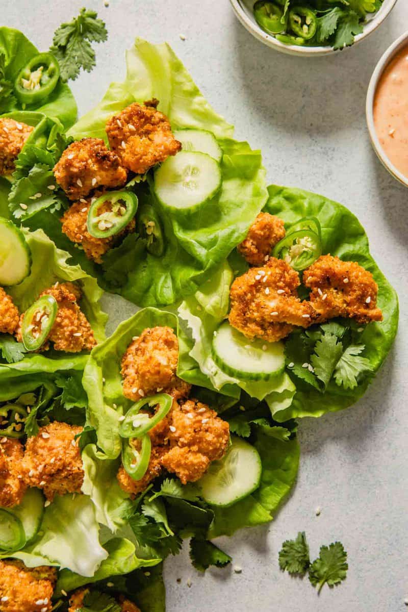 Overhead image of lettuce wraps stacked up along eachother on a light blue table. Lettuce wraps are filled with coated cauliflower, cucumbers, serranos and sesame seeds.