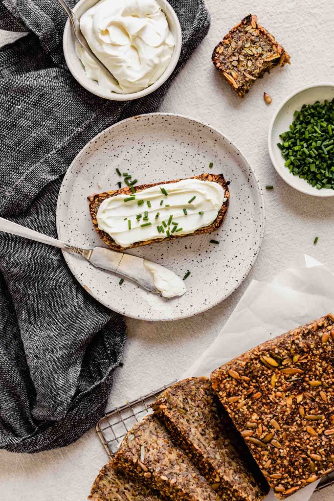 Overhead image of nut and seed bread set on a table with plates of bread slices topped with cream cheese and chives