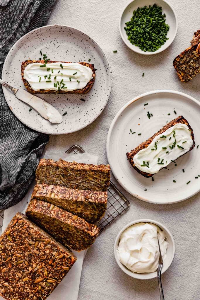 Overhead image of nut and seed bread set on a table with plates of bread slices topped with cream cheese and chives
