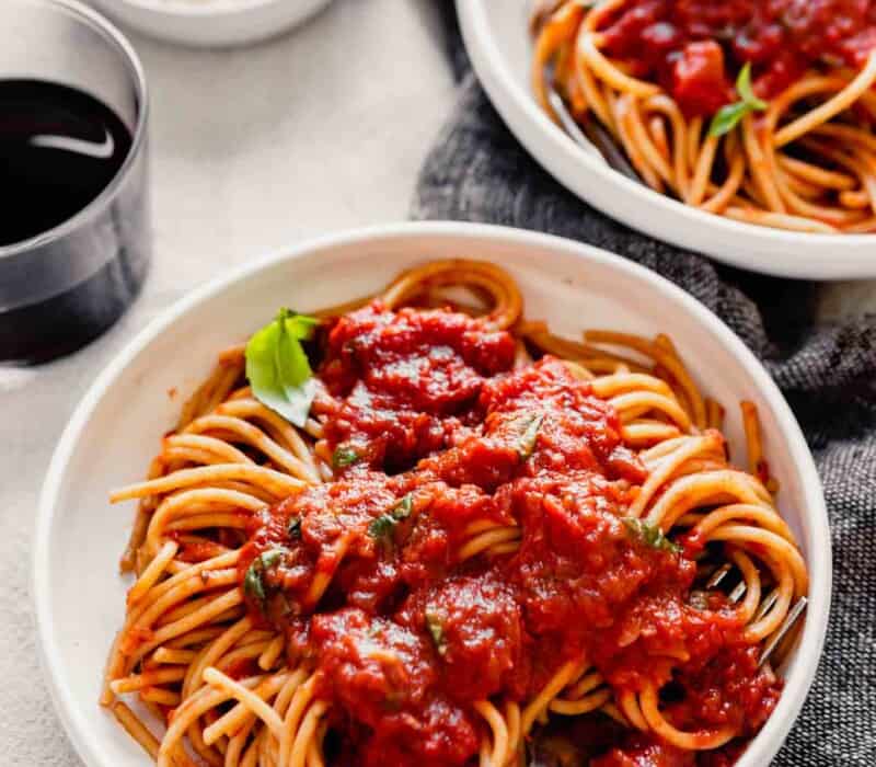 side angle of spaghetti and red sauce in a white bowl on a table with wine and cheese
