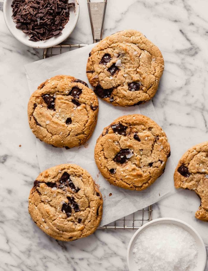 Overhead image of chocolate chip cookies stacked on a cooling rack on top of a marble table with bowls of salt and chocolate set around