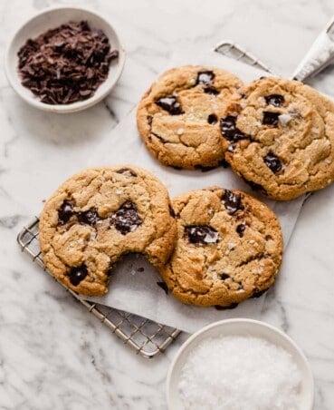 Overhead image of chocolate chip cookies stacked on a cooling rack on top of a marble table with bowls of salt and chocolate set around
