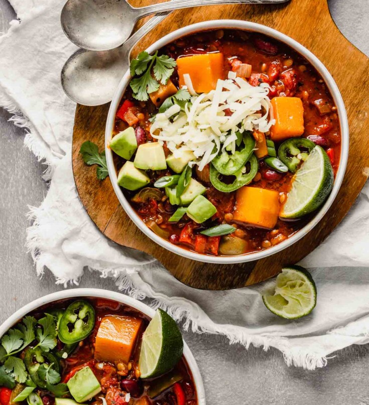 Overhead image of chili in two white bowls, one set on a wood board, the other on the counter. Chili bowls topped with avocado, cilantro, jalapenos, cheese. Lime wedges scattered around.
