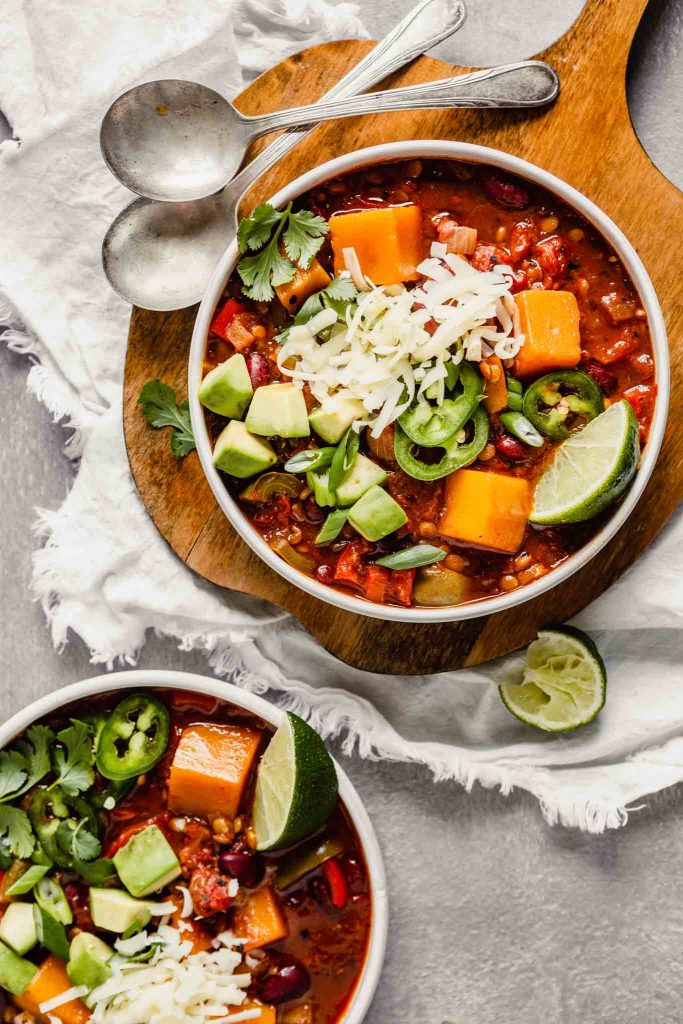 Overhead image of chili in two white bowls, one set on a wood board, the other on the counter. Chili bowls topped with avocado, cilantro, jalapenos, cheese. Lime wedges scattered around.