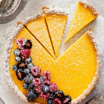 Overhead image of a lemon tart set on parchment paper with powdered sugar and berries scattered around it