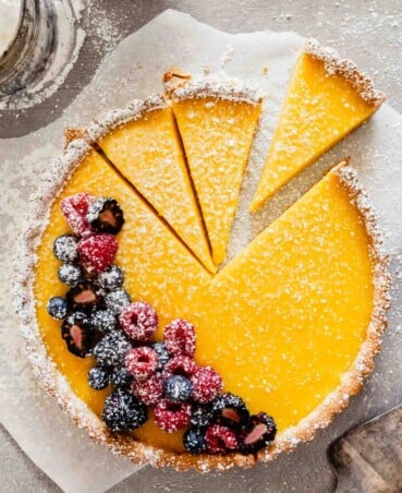 Overhead image of a lemon tart set on parchment paper with powdered sugar and berries scattered around it