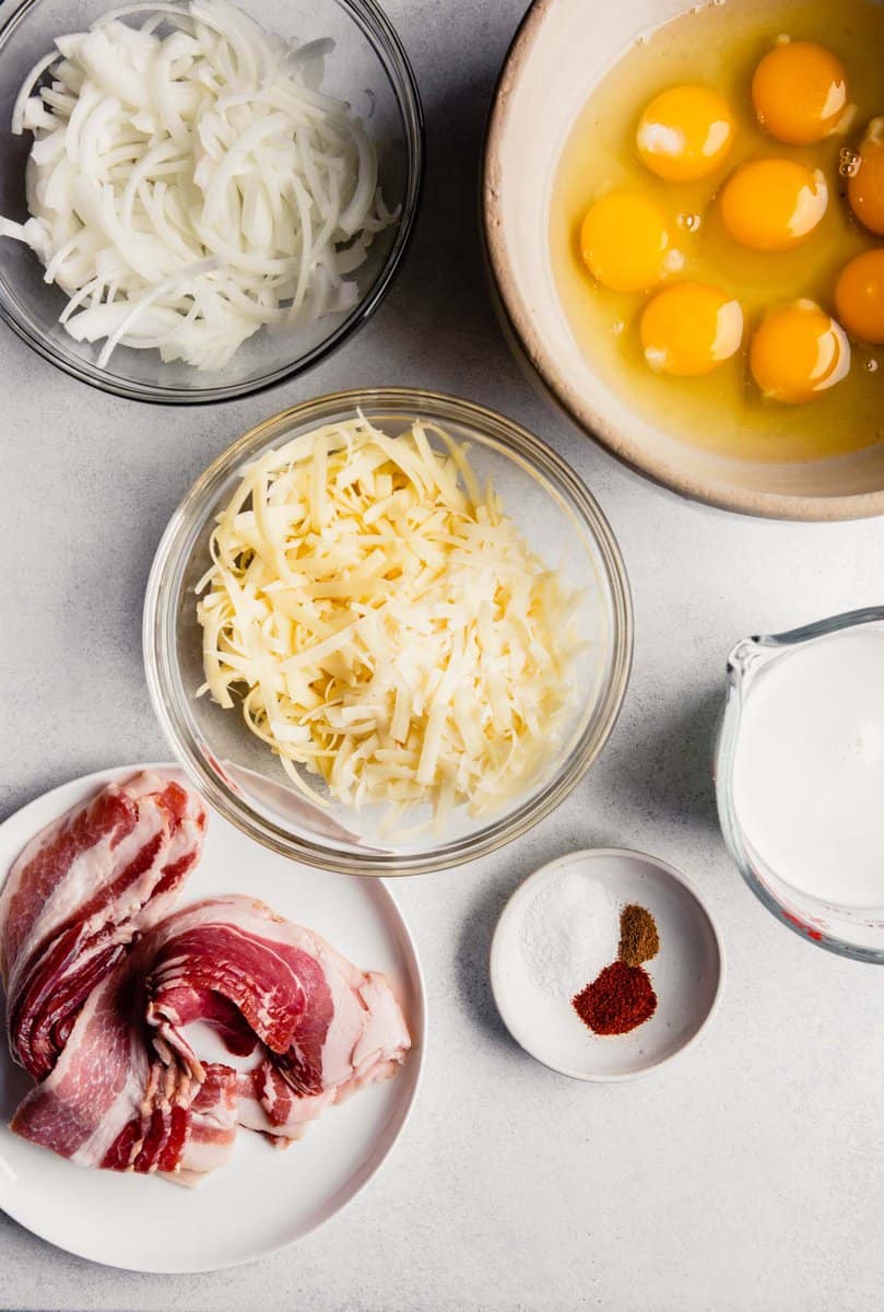 Overhead image of eggs, cheese, onion, bacon, milk and spices prepped and set on a table 